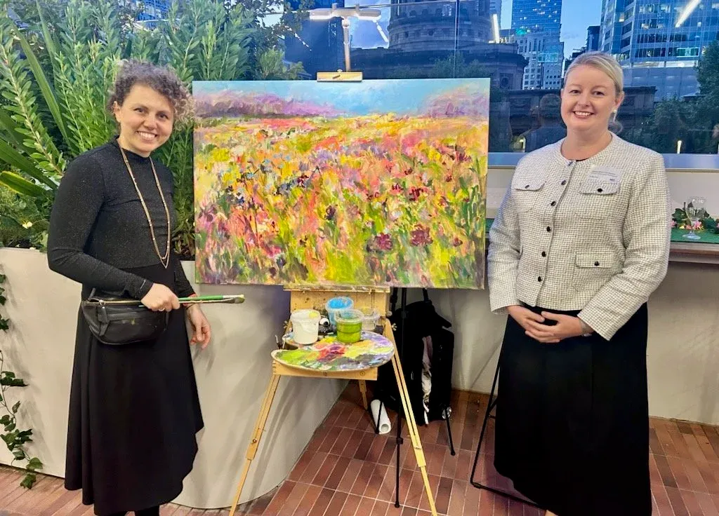 Two women standing next to an easel with a colorful landscape painting inside, surrounded by green plants, in an indoor setting with city buildings visible through the windows.