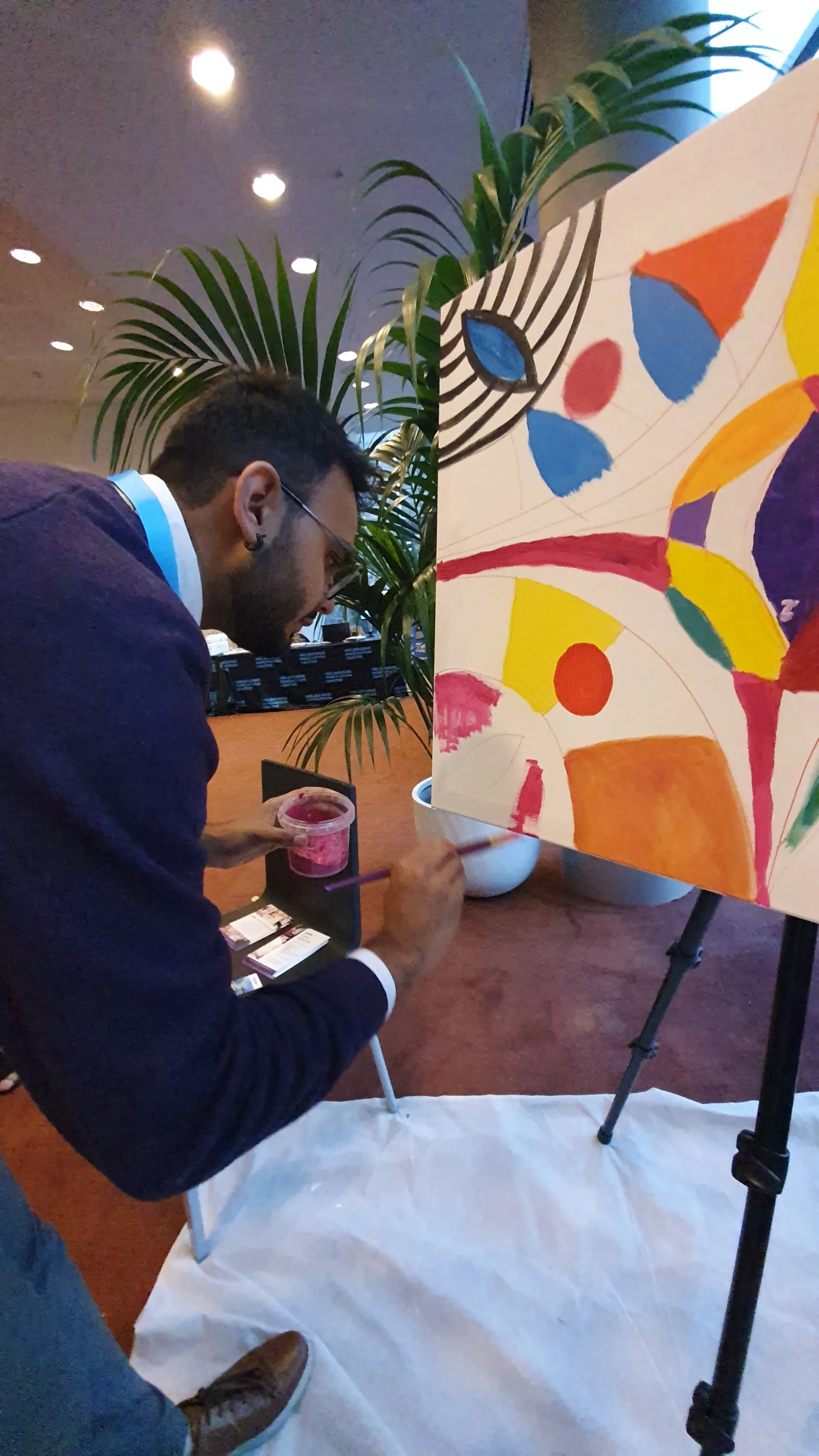 A man painting an abstract artwork on a large canvas at an indoor event. He is holding a cup of pink paint and using a brush. The event space has a large green plant and ceiling lights visible in the background.
