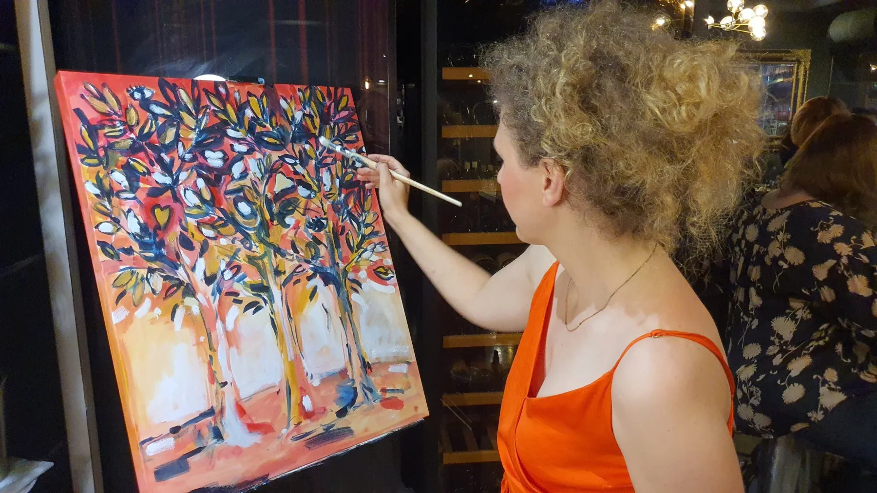 Woman with curly hair painting an abstract colorful tree on a canvas at an indoor art event.