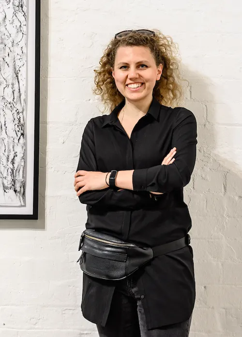 A woman with curly hair smiling and crossing her arms, standing against a white brick wall.