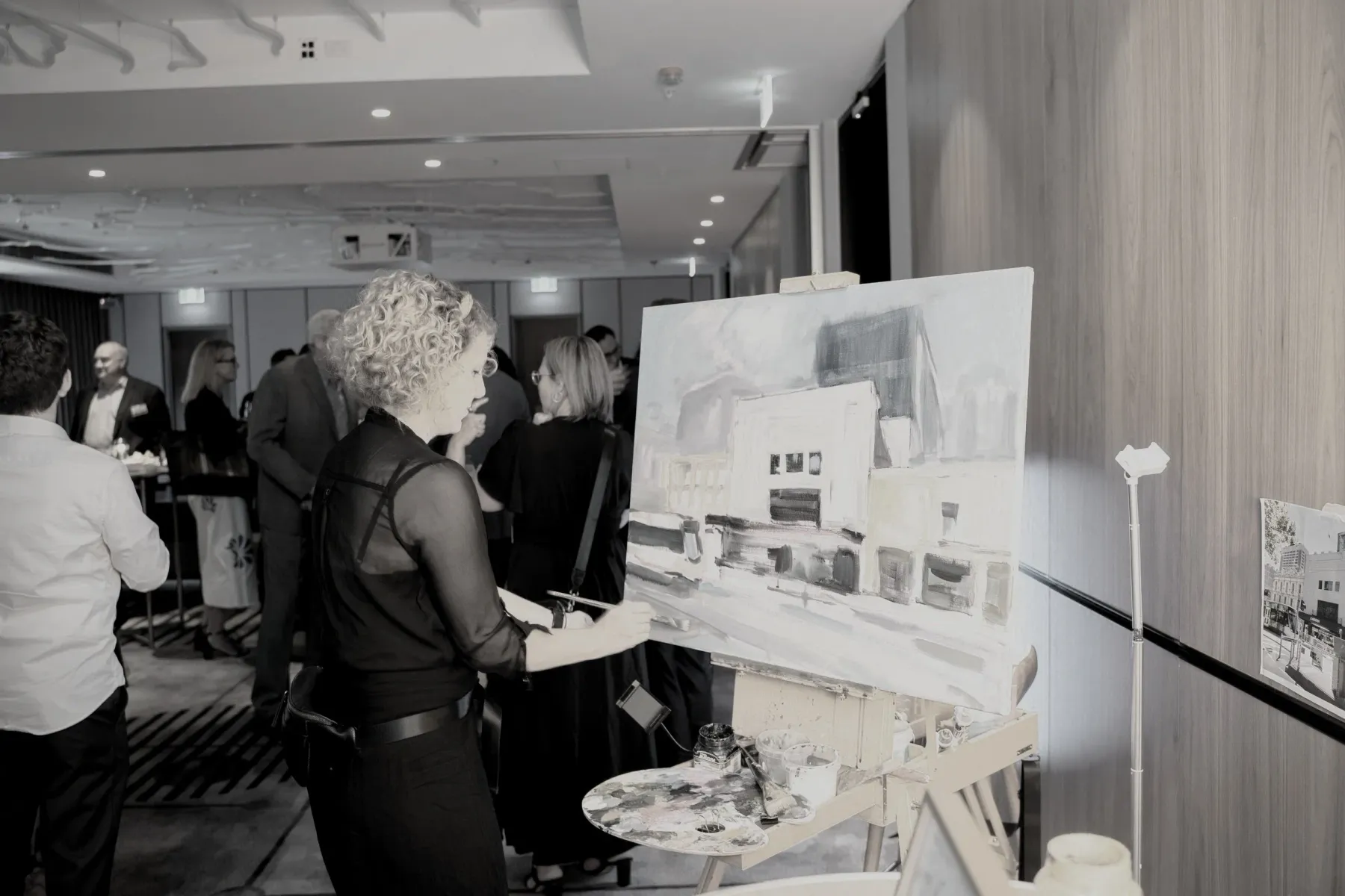 A woman with curly blonde hair painting a landscape on an easel at an event. She is wearing a black sheer top and stands in front of the artwork. Several other people are present in the background, mingling and viewing art.