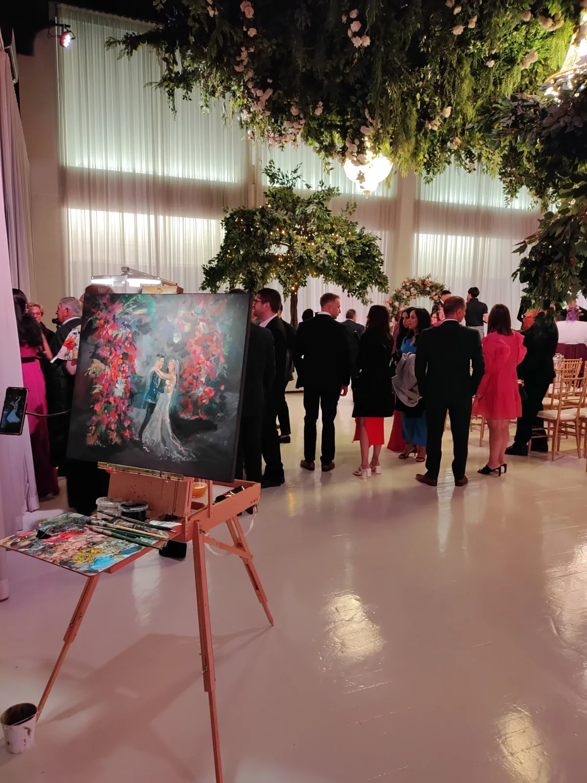 An indoor event with people dressed for a formal occasion, artwork on display on an easel, a tree with lights, and floral decorations, under a ceiling of large curtains and a chandelier.