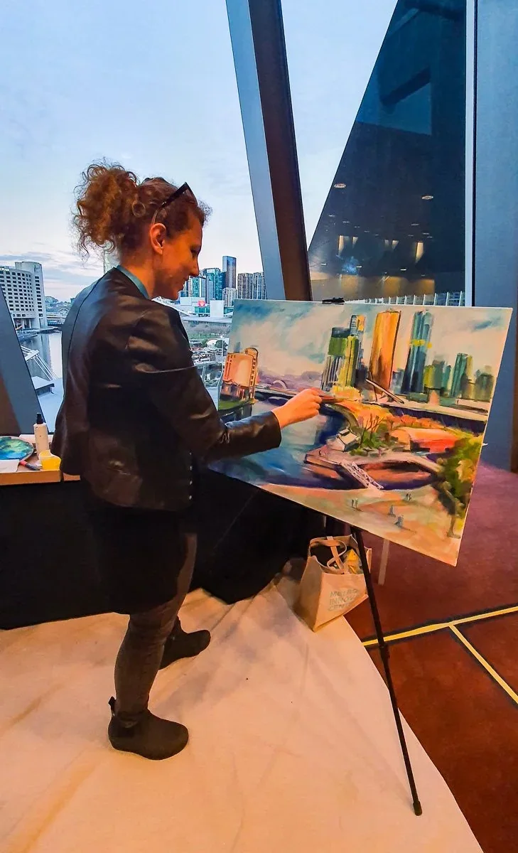 A woman with curly red hair in a bun, wearing a black leather jacket and black pants, is painting a cityscape on a canvas. She is standing indoors near large windows overlooking a city skyline with tall buildings.