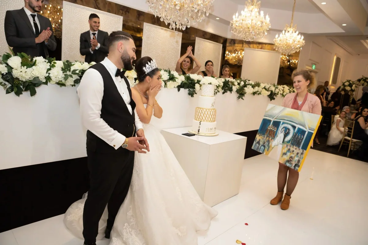 A wedding reception with the bride and groom standing in front of a three-tiered wedding cake decorated with gold accents. The bride is wearing a wedding gown and tiara, and the groom is in a tuxedo. Guests are clapping and smiling, with women in bla