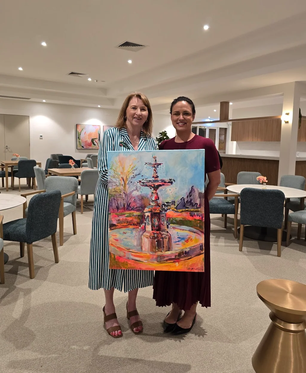 Two women standing at Fountain Court holding a colorful painting of a fountain with trees and mountains in the background.