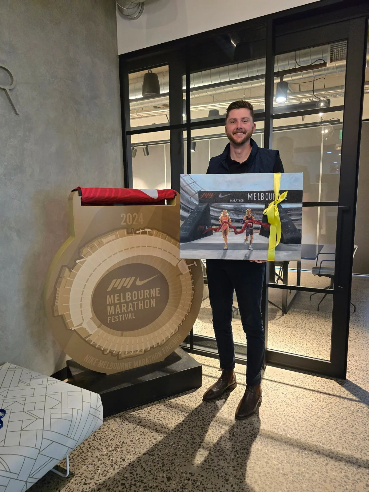 A man with short brown hair, beard, dark shirt, navy vest, and brown boots smiling while standing inside a modern indoor venue, holding a Nike Melbourne Marathon Festival painting, with a large gold medal display and a window reflecting industrial ce