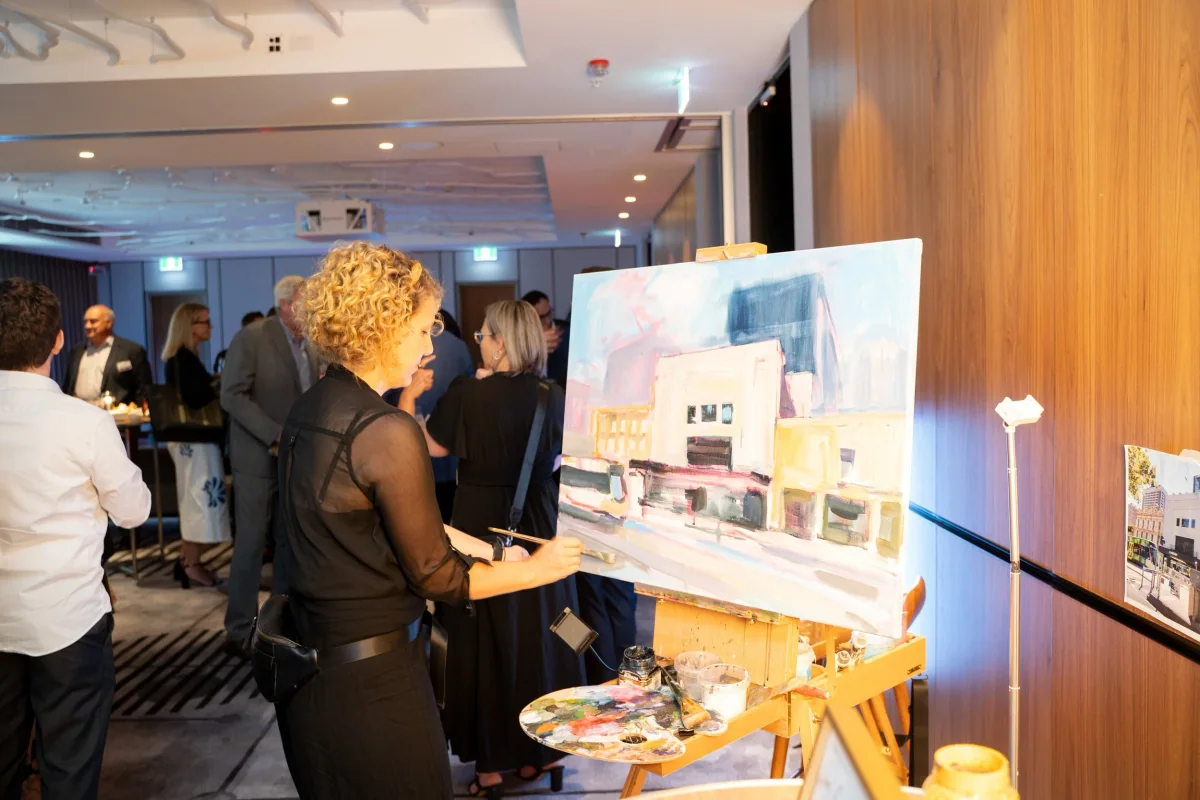 A woman painting a cityscape on a canvas at an AHA hospitality event with several people socializing in the background.